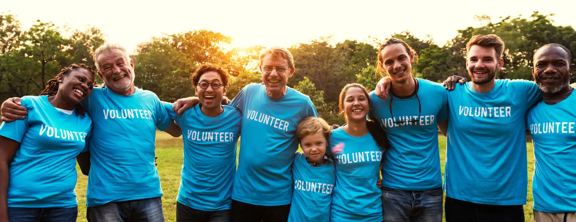 A group of charity volunteers in a field of all ages stand with arms over shoulders and smile at the viewer